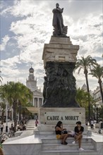 Large statue with people, surrounded by palm trees and historic architecture, Cadiz