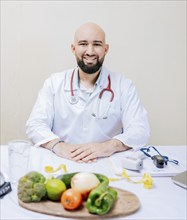 Smiling nutritionist doctor at desk with laptop and vegetables. Bearded nutritionist doctor at his