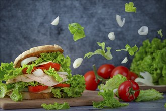 Sandwich on a wooden board with tomatoes, lettuce, sausage, onion in front of a dark background