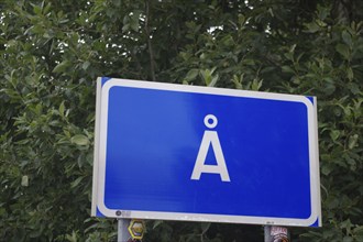 Blue road sign with white frame and the letter Å against a background of dense green foliage, the