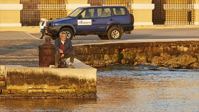 A man relaxes while fishing by the sea next to a parked off-road vehicle, Mandraki Oat, harbour