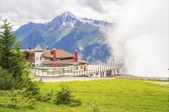 A building on a hill in front of a mountain backdrop with clouds and a green meadow, quiet and