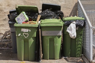 Green and grey plastic recycling and thrash bins filled with cardboard and black garbage bags in a