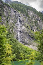 A high waterfall cascades down a rocky mountain face, surrounded by green trees and lush nature,