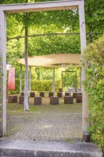 Wooden frame and stacked wooden cubes under a shady area in a garden or park, surrounded by plants,