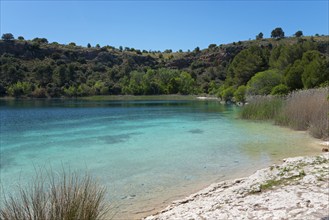 A calm lake with clear blue water, surrounded by green trees and rolling hills under a clear sky,