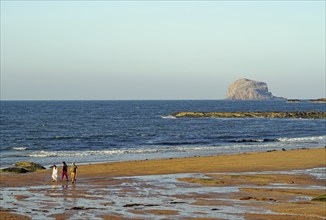 Walkers on the beach with rocks and waves in the background under a clear sky, Bass Rock, North