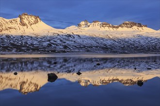 Snow-covered mountains reflected in a fjord in the evening light, Berufjördur, East Fjords,