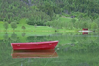 A small red boat floats on a calm lake with green surroundings, Stryn, Mindesundet, Fjordland,