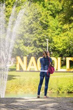 A woman photographed in front of a fountain in a sunny green park with trees in the background,