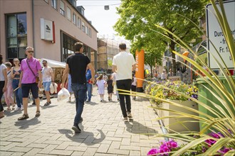 Lively street scene with people walking, surrounded by shops and flowers in a summer atmosphere,