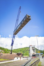 A crane lifts a bridge over a building complex in sunny weather with blue sky and white clouds,