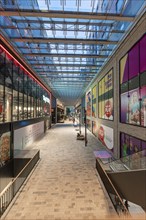 Modern shopping centre with glass roof, brightly lit and lined with shops. Colourful advertising