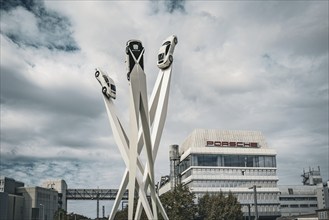 Modern sculpture of cars on high pillars in front of a modern building under a cloudy sky,