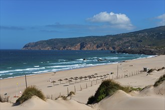 Beach with sand dunes and sunshades, behind them the sea and rocks, partly cloudy sky, moving dune,