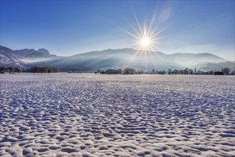 Winter landscape with snow-covered field and rising sun against a mountain backdrop, Bad Feilnbach