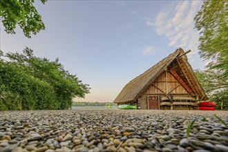 Thatched roof house next to a gravel path, surrounded by trees and grass under a cloudy sky, with