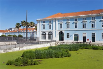 A large blue building with many windows and a green pond in the foreground under a sunny sky,