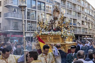 A traditionally decorated procession passes by buildings, cheered on by spectators. The atmosphere