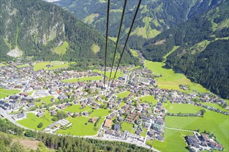 View from above of a town with surrounding mountains and green meadows, with cable car cable in the