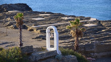 View of a rocky coastline with palm trees, a white bell frame and blue sky in the background, rock