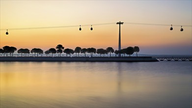 Silhouettes of trees and cable cars against a serene sunset reflecting on the calm water, Lisbon