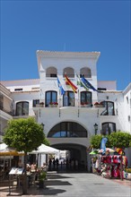 City gate of a whitewashed building on a square with flags and shops under a clear blue sky, town