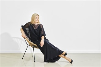 Calm and positive woman in black shiny dress lounging on a modern wicker chair in a bright room