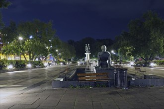 City square illuminated at night with fountain, statues and surrounded by trees and benches, square