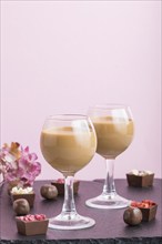 Sweet chocolate liqueur in glass with chocolate candies on a pink background and black stone slate