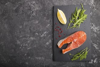 Raw salmon steak with arugula and lemon on a black slate board on a black concrete background. Top