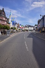 Main street with bus stop Adenau Markt, buildings, half-timbered houses, flags and trees under blue