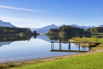 A quiet lake with a jetty, surrounded by trees and mountains under a clear blue sky, Forggensee