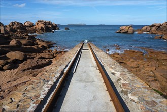 A stone path leads through a rocky coastal landscape to the blue sea under a clear sky, Cale de