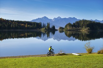 A cyclist rides along the shore of a lake, surrounded by mountains and reflecting trees in a clear