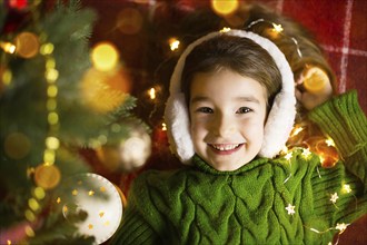 A happy girl in fur headphones lies near the Christmas tree in garlands and lights in a warm