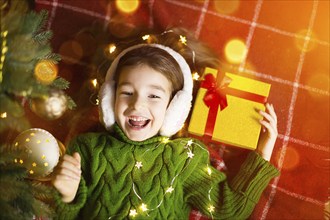 Girl in a warm knitted sweater and fur headphones is lying on a blanket near the Christmas tree