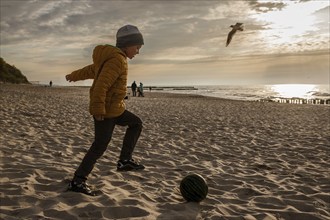 Boy playing football on the beach by the sea during sunset.