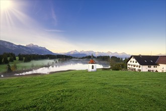 Farmhouse next to a chapel on a lake with a view of the Alps at sunrise, surrounded by green