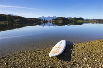 A calm lake with a surfboard on the shore, surrounded by mountains and a clear blue sky, Forggensee