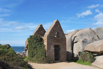 Stone ruin on the coast with sea view, surrounded by rocks and light vegetation under a blue sky,