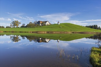 View of a lake with reflection of a house and a chapel on a hill, Hegratsrieder See near Füssen,