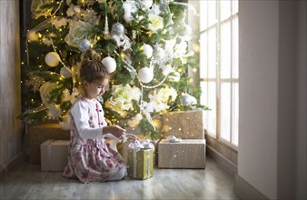 Little girl in beautiful dress is sitting under Christmas tree with gift box and bow. Light from a