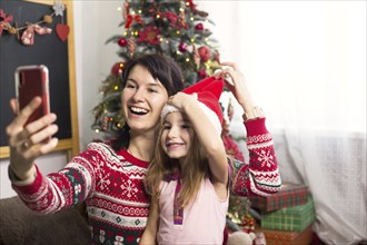 Mom and daughter are sitting near the Christmas tree, hugging, taking photos and selfies on the