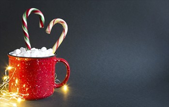 Red mug with marshmallows and caramel candy cane in the shape of a heart and lights of garlands on