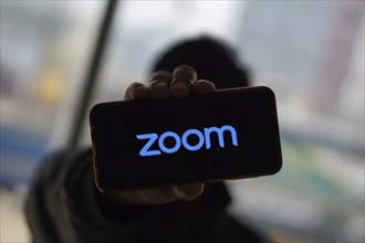 Zoom App logo on smartphone screen while Zoom meeting in the background.