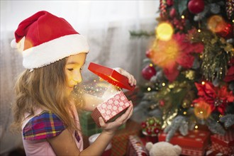 Little girl in Santa hat opens a red box with a gift and a Golden magic light near the Christmas