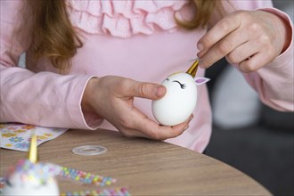 A cute girl with pink bunny ears makes an Easter craft, decorates an egg in the form of a unicorn