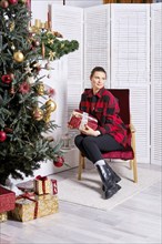 Pensive woman in warm plaid shirt and tight trousers holds christmas gift box in hand and looks to