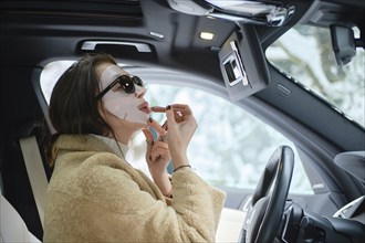 Concept of the rapid pace of life, woman applying lipstick wearing face mask while driving a car